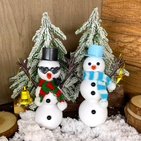 Two decorated snowmen sit in front of flocked evergreen trees. 